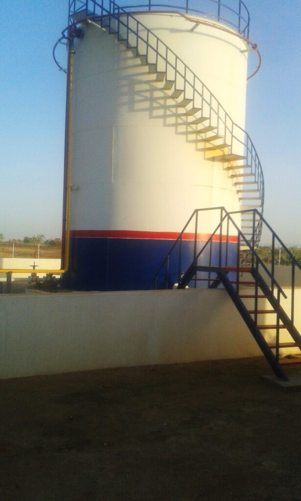 A vertical storage tank for petroleum derivatives, equipped with a spiral staircase for technical access and a concrete detention basin (Bund Wall) to ensure environmental safety.