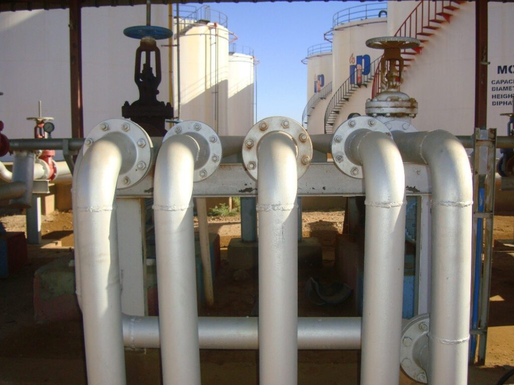 The transportation and transformation pipeline manifold system is equipped with manual gate valves to control the flow of petroleum derivatives, and vertical storage tanks with spiral staircases appear in the background to ensure the smooth flow of operational processes.