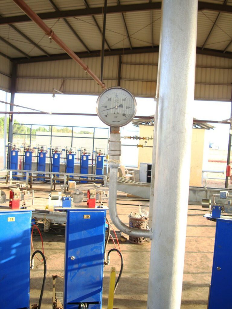 A pressure gauge is mounted on a main pipeline inside the processing unit, used to monitor operational pressure and ensure it remains within safe limits, with rotary filling platforms appearing in the background.